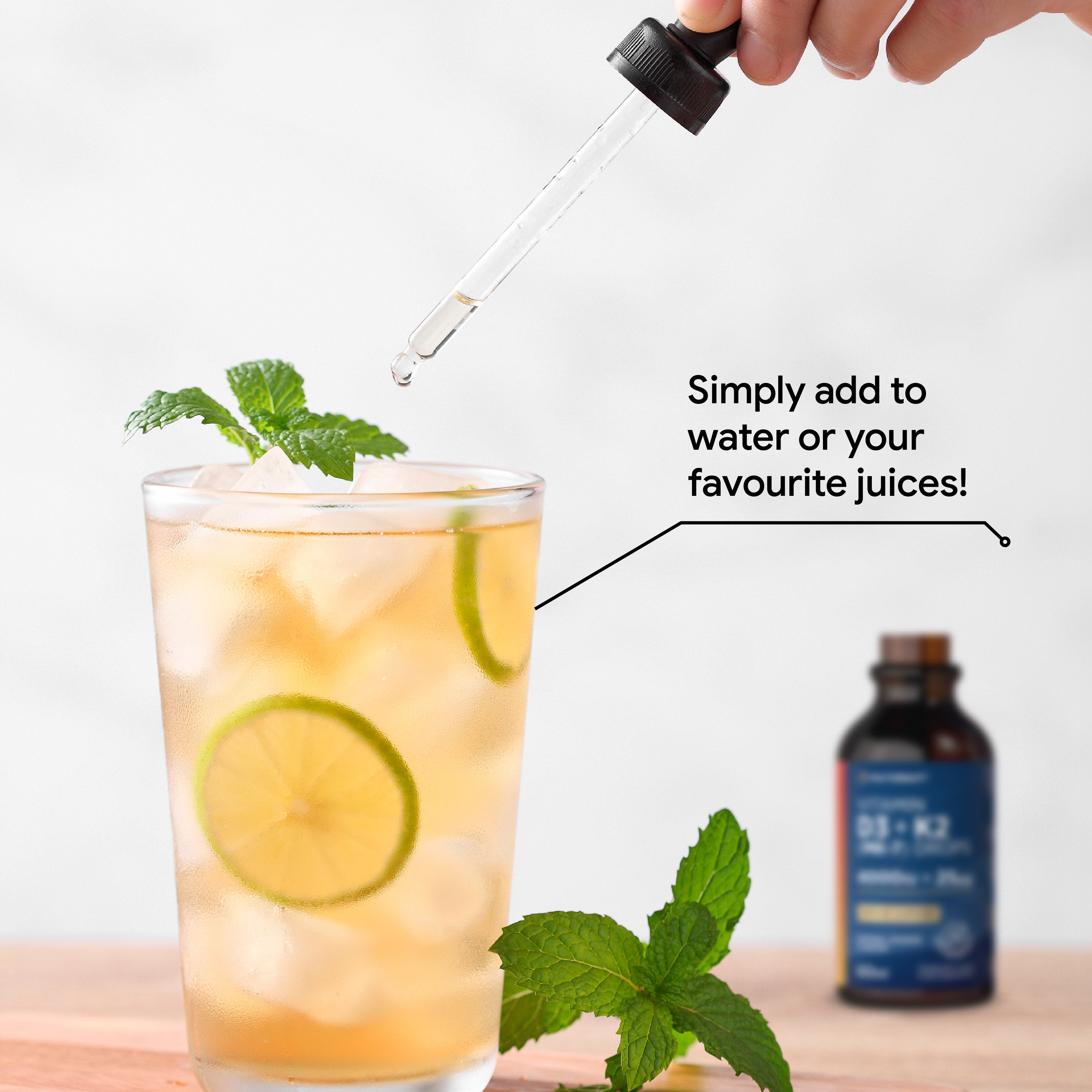 A hand using a dropper to add Vitamin D3 & K2 liquid into iced juice with lime and mint.