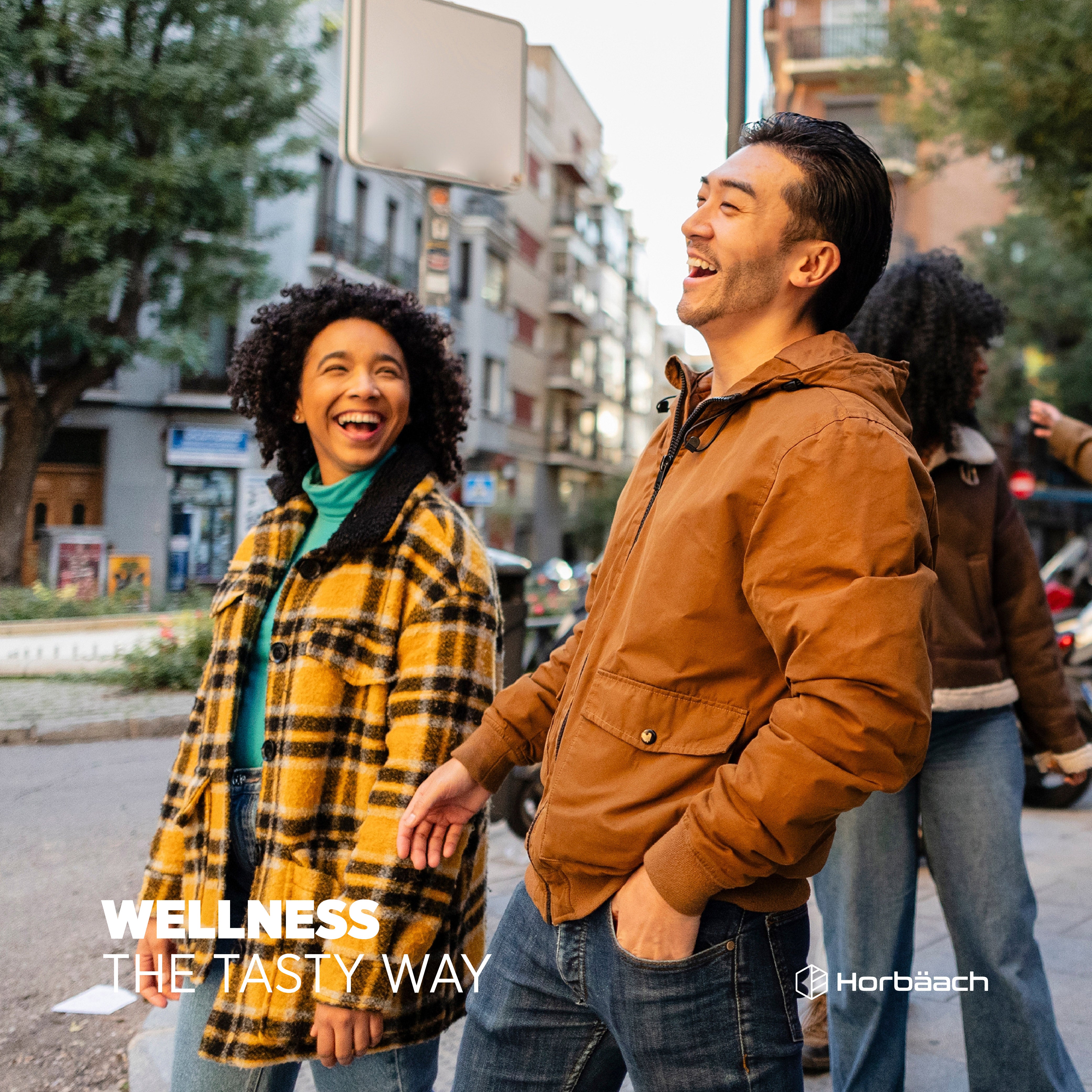 Young adults laughing and walking on a city street, with the text 'WELLNESS THE TASTY WAY' and Horbäach logo visible.