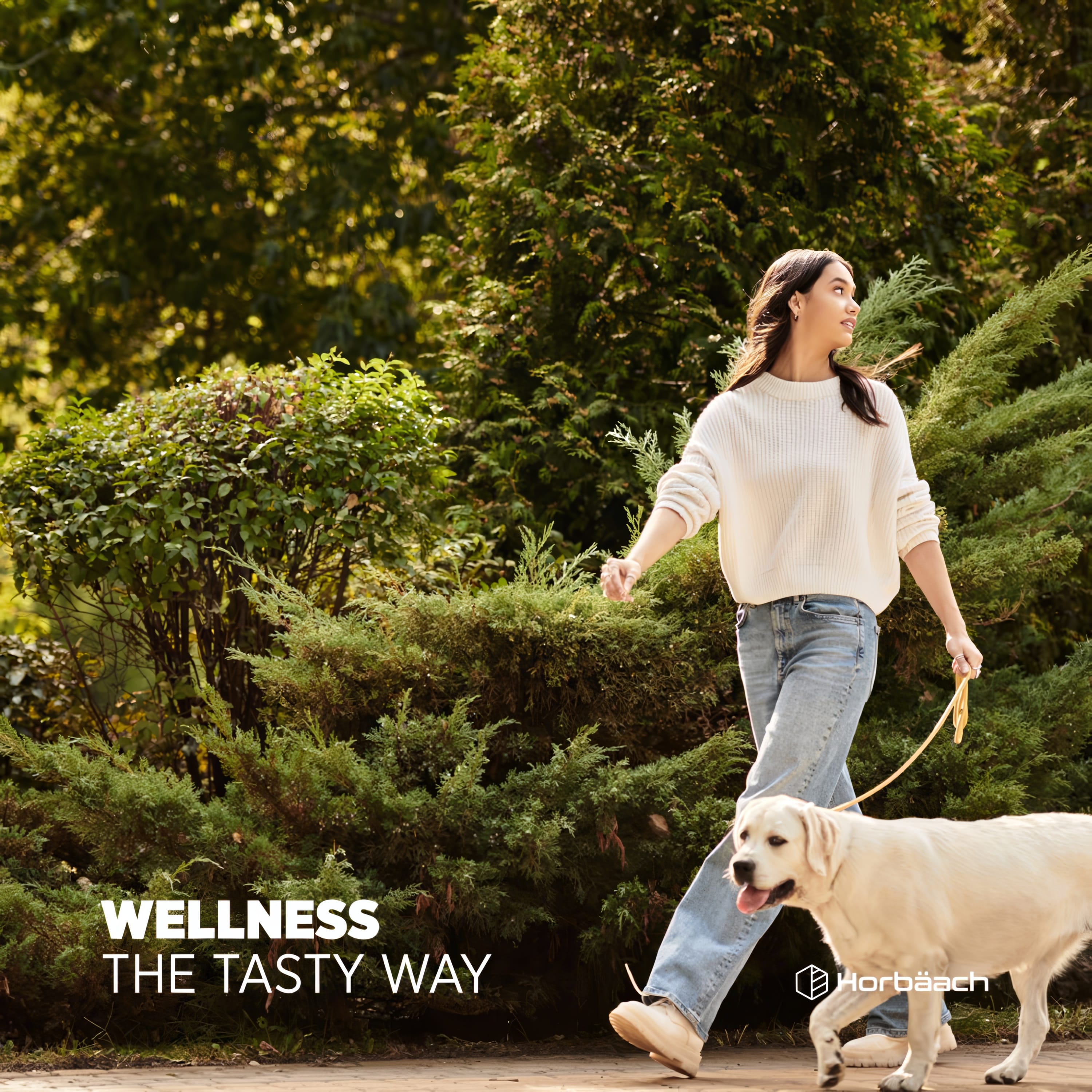 Young adult woman walking a golden retriever on a paved path in a sunny park with green bushes and trees.