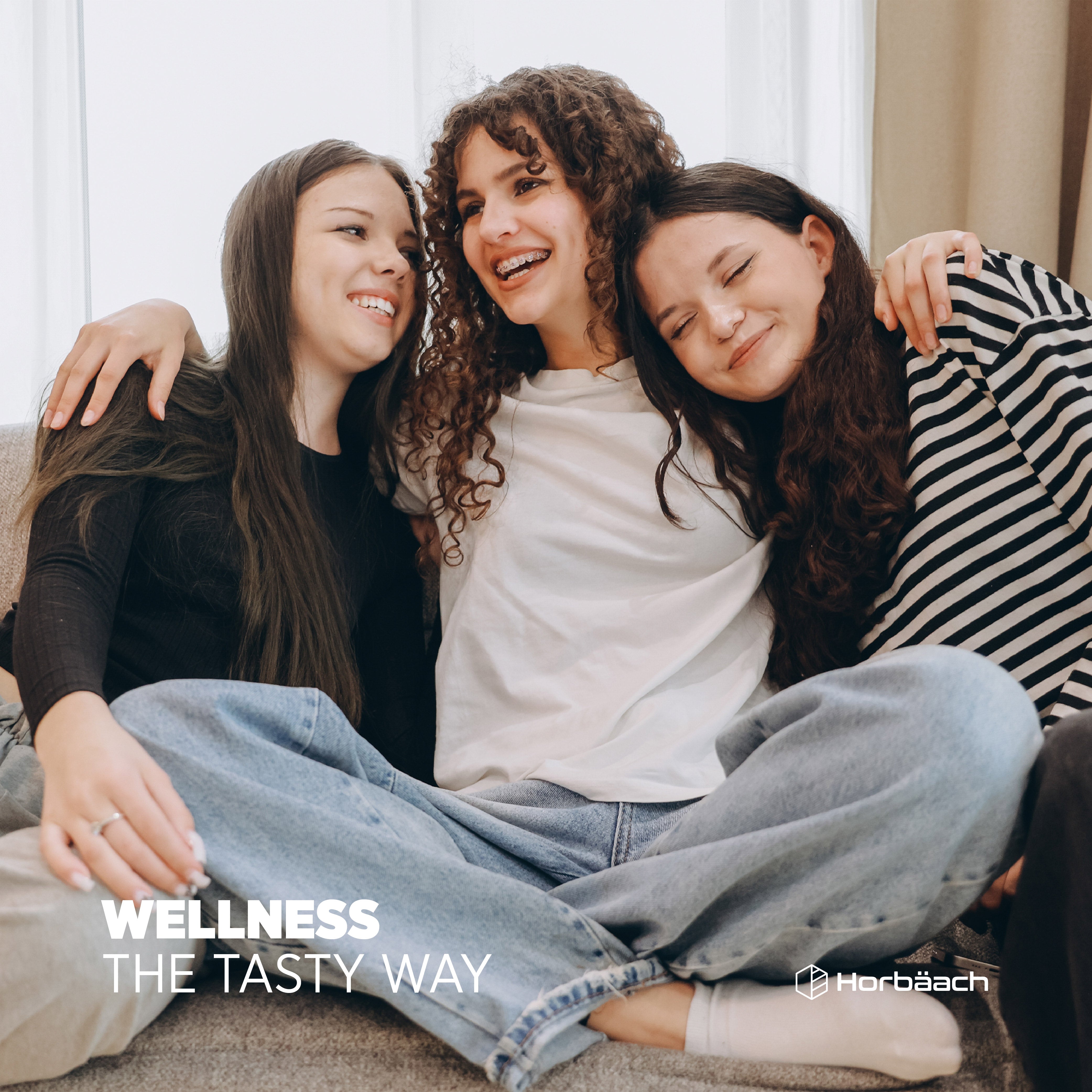 Three young women smiling and hugging on a couch. Text reads 'WELLNESS THE TASTY WAY' with 'Horbäach' logo visible.